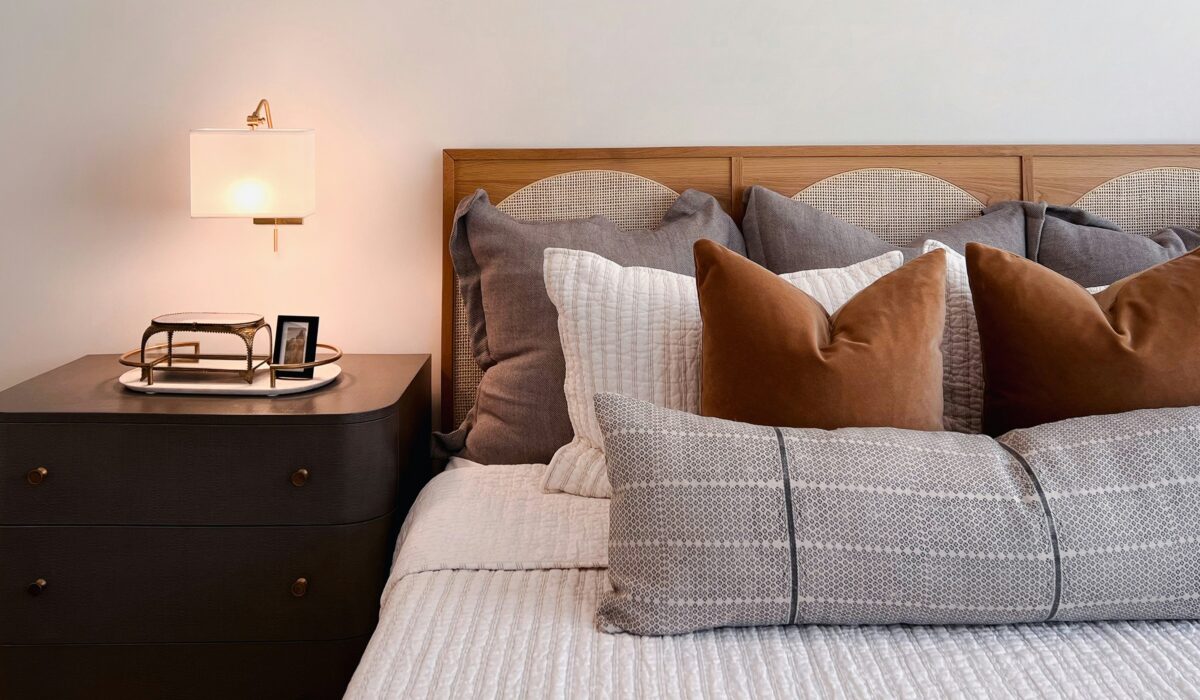 modern bedroom with wooden headboard, grey bedding, and brown accent cushions beside dark bedside table with small lamp and book