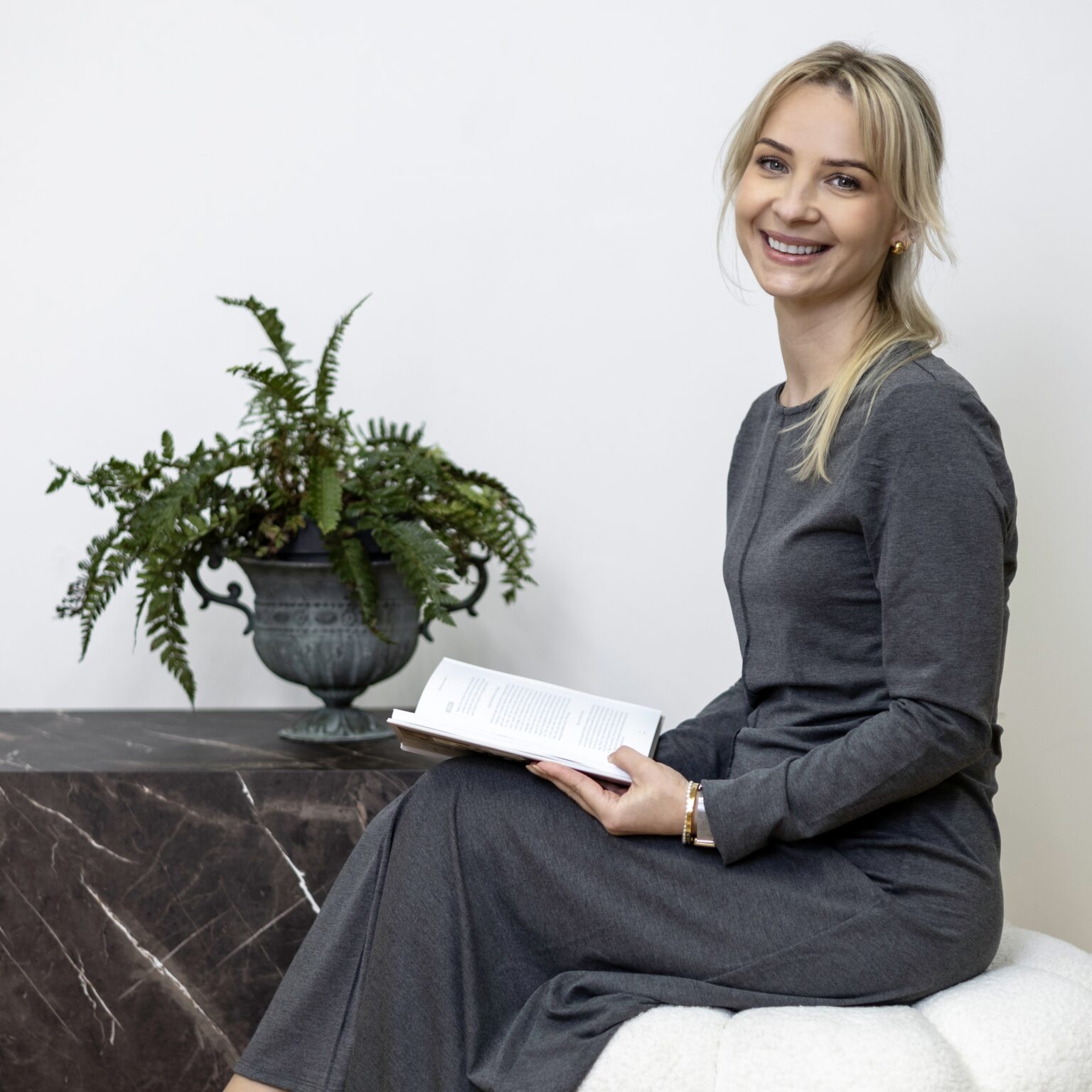 Monika Clayton, South London interior designer, seated on a white boucle pouffe holding a book in a calm interior setting